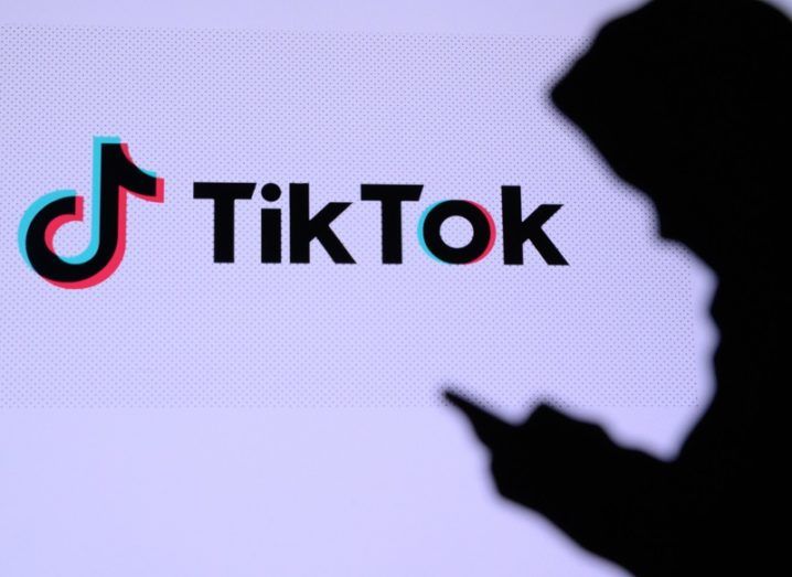 Silhouette of person holding a phone beside the TikTok logo on a white wall.