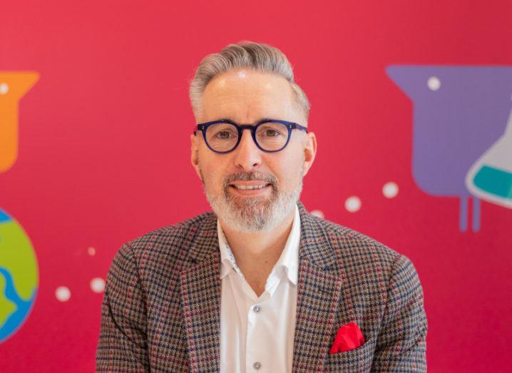 Martin McKay wears dark round-rimmed glasses and a grey suit with a red pocket square, sitting against a bright red background.