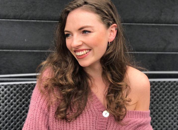 Sarah Dillon wearing a pink jumper smiling in front of a grey brick wall.