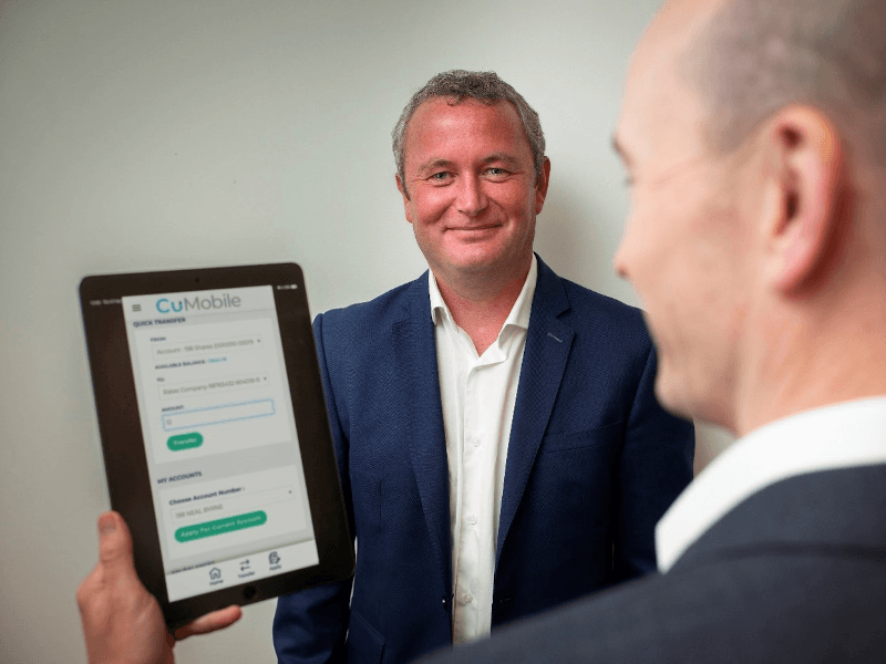 Two men standing in a white room, one is holding an iPad with the new CuMobile app displayed on its screen.