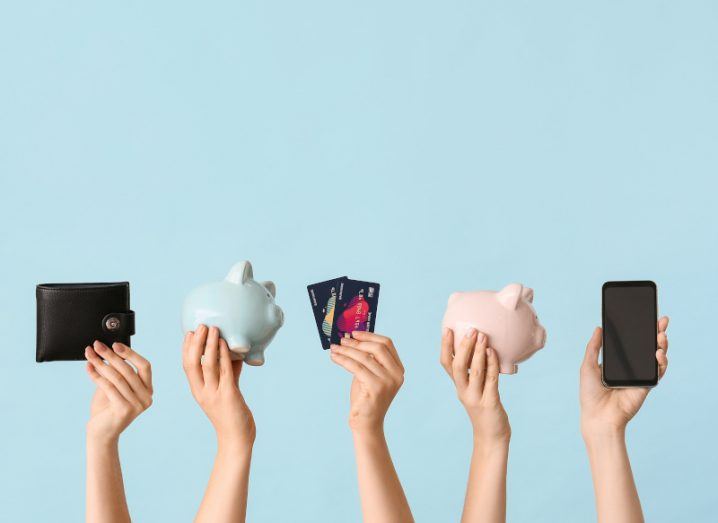 Five arms come up from the bottom of the shot, each holding one item; a wallet, a blue piggy bank, a pair of credit cards, a pink piggy bank and a phone.