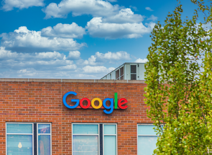 The Google logo on the side of a red brick building. There is a tree in front of the building.