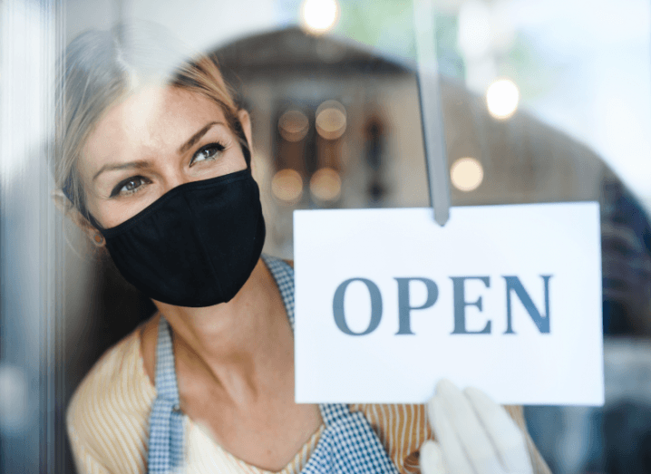 A person wearing a black mask holds an open sign in a shopfront.