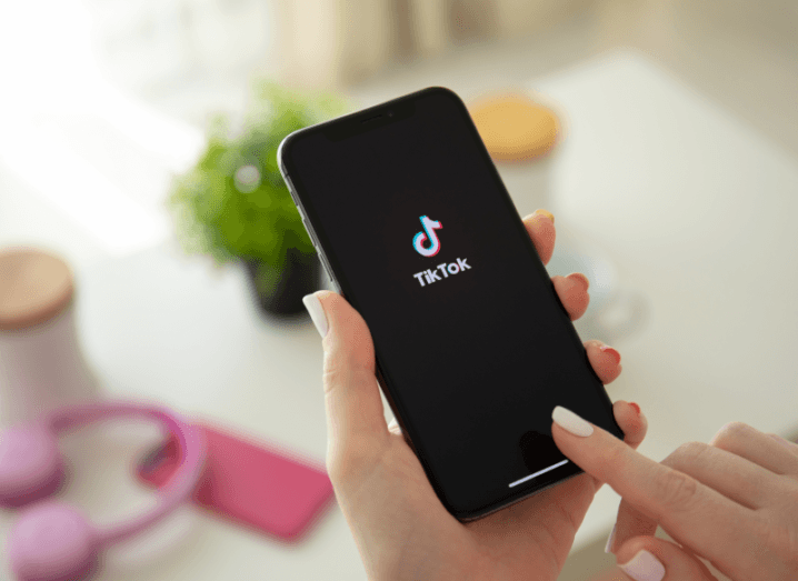 The TikTok app displayed on the screen of an iPhone. In the background there are headphones and a potted plant on a table.