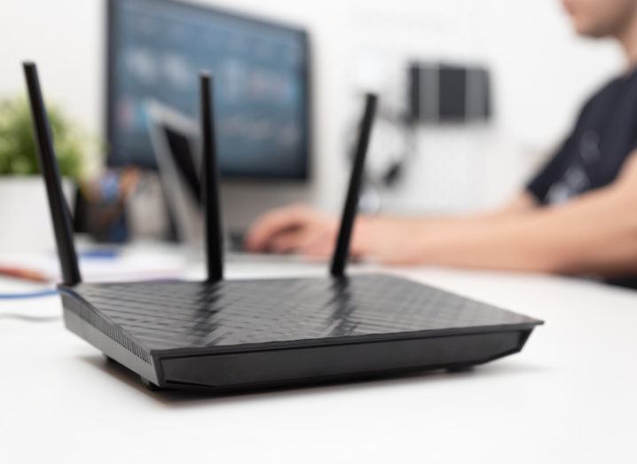A black wireless broadband router is sitting on a desk, while a man is working on a computer in the background.
