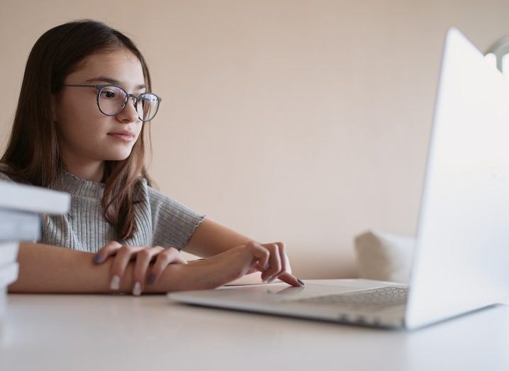 Young girl wearing glasses at a laptop against a white wall background.