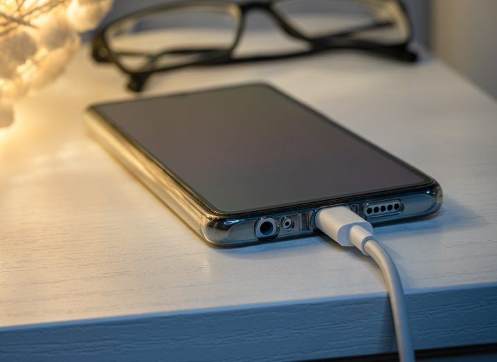 Smartphone charging at night on a bedside table with a pair of glasses beside it.