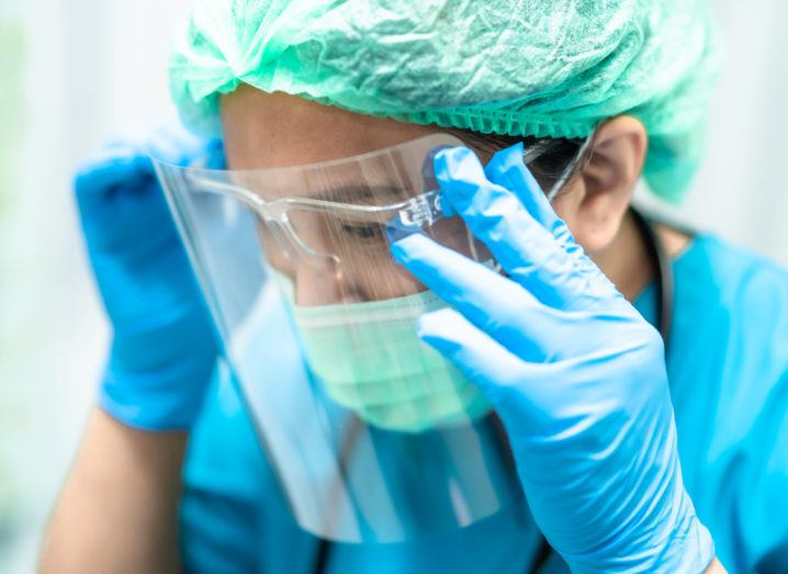 A woman holds her hands up to adjust a face shield. She is wearing blue disposable gloves, goggles, green hair covering and face mask, and blue scrubs.