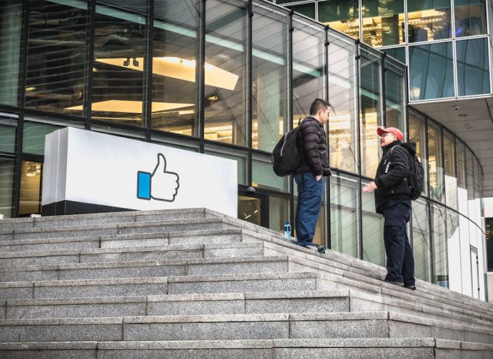 Two people stand on the stone steps leading to a glass-fronted office building. A sign next to them bears the Facebook thumbs-up icon.