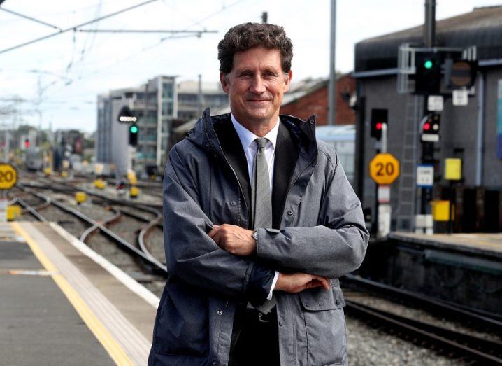Eamon Ryan in a grey jacket with folded arms in front of train tracks.