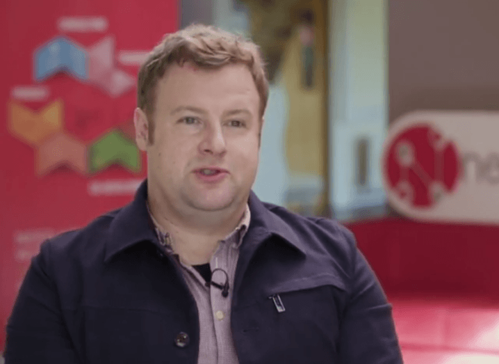 A man speaks to camera in front of a NearForm logo in an office with cherry-red accents.