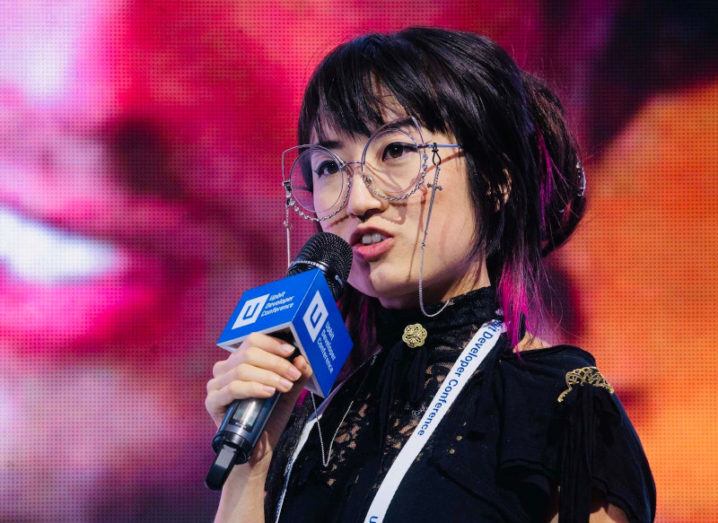 A close-up of Yan Zhu, a young woman wearing large glasses speaking at a conference while holding a microphone.