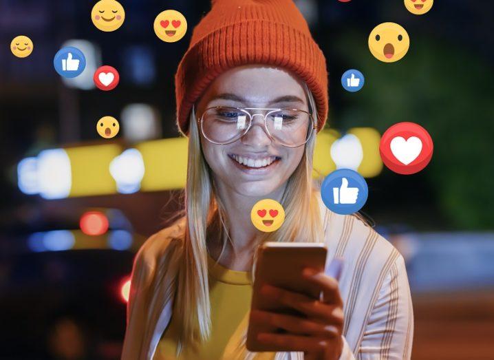 Woman wearing orange beanie hat and glasses smiling while looking at her phone surrounded by heart and thumbs-up emojis.