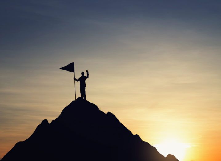 A business person standing on top of a mountain at sunset, planting a flag into the peak.