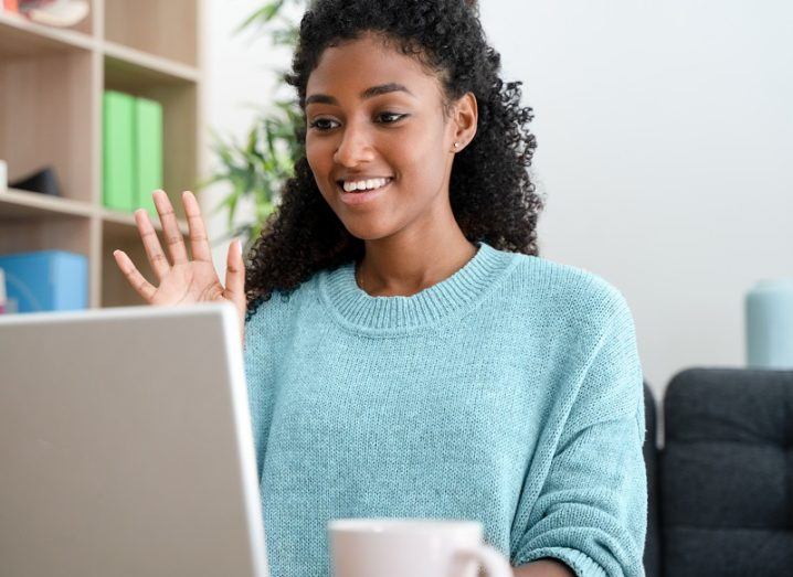 Woman wearing a blue jumper waving at a laptop screen on a Zoom call.