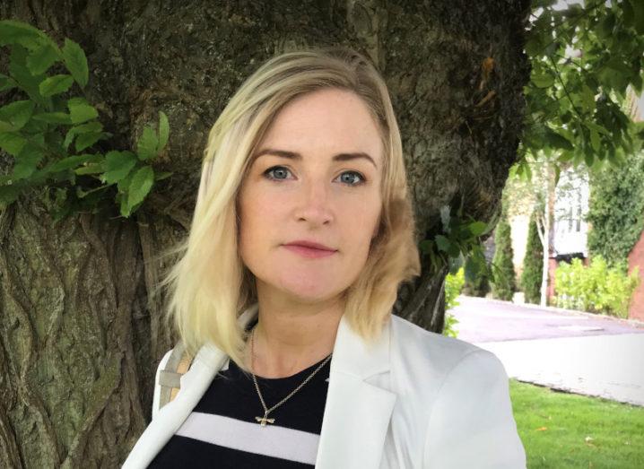 A woman with blonde hair wearing a white blazer stands outside in front of a leafy tree. She is smiling at the camera.