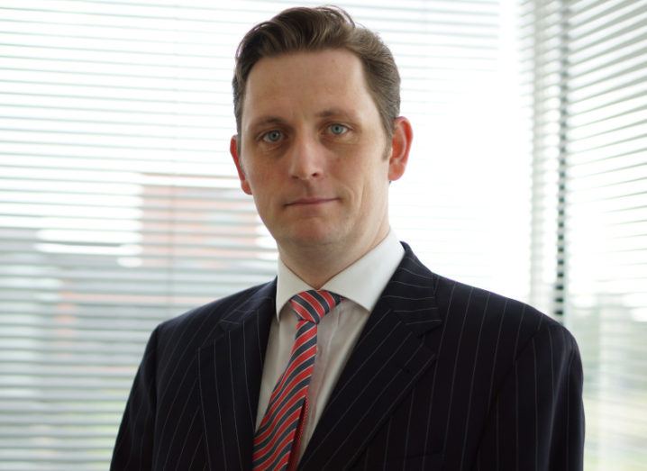 Sean Keating is standing in front of an office window wearing a suit.