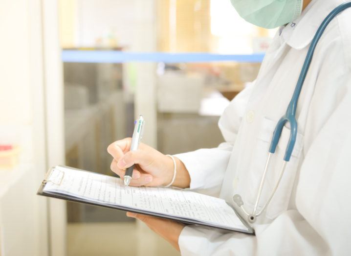 A scientist is standing in a lab and writing on a document on a clipboard while wearing a labcoat and surgical mask.