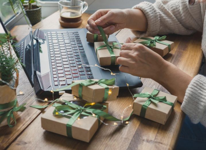 A woman’s hands tie small brown-paper packages with green ribbon with a laptop open nearby for online shopping.