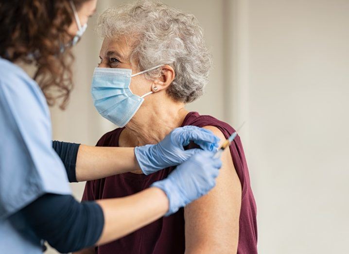 Elderly woman wearing a face mask and receiving a vaccine from a doctor in blue scrubs.