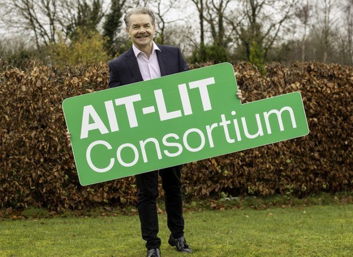 Prof Ciaran O'Cathain smiling in a suit, holding a green sign that says AIT-LIT Consortium.