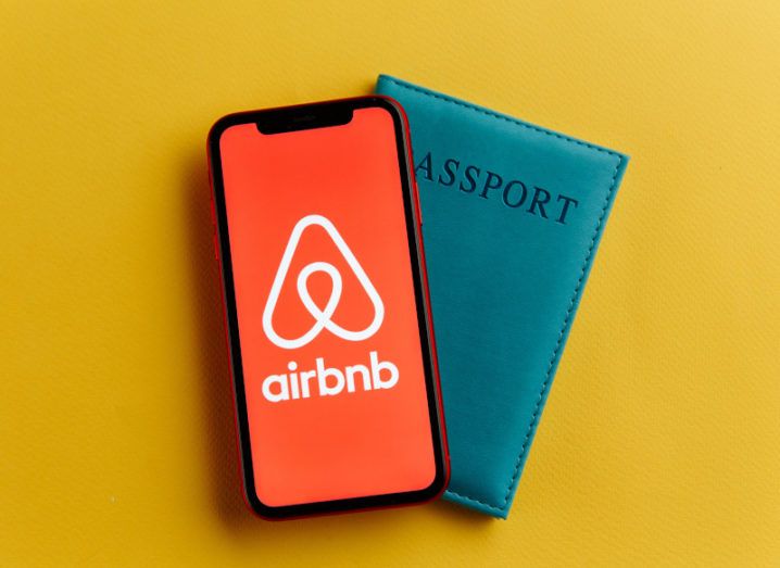 A black smartphone with a full-screen picture of the Airbnb logo on it. The phone sits on top of a dark green passport against a yellow background.