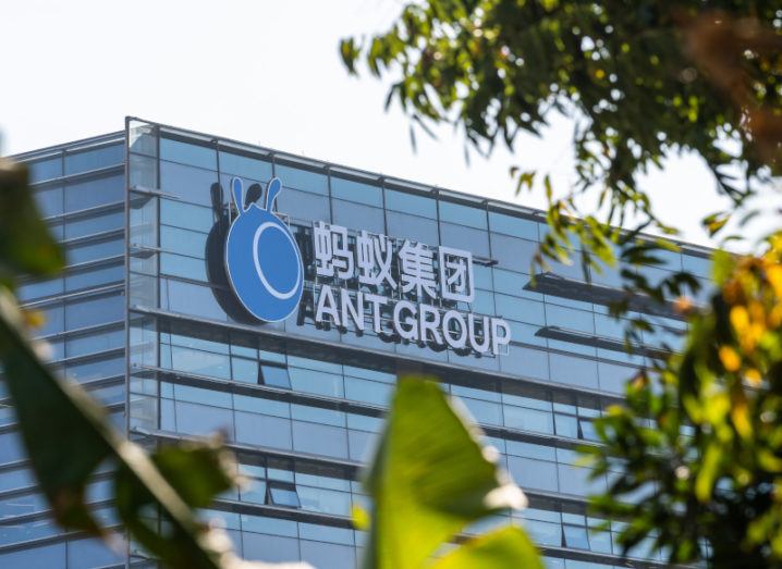 A shot through some greenery of the Ant Group logo on top of an office building against a blue sky.