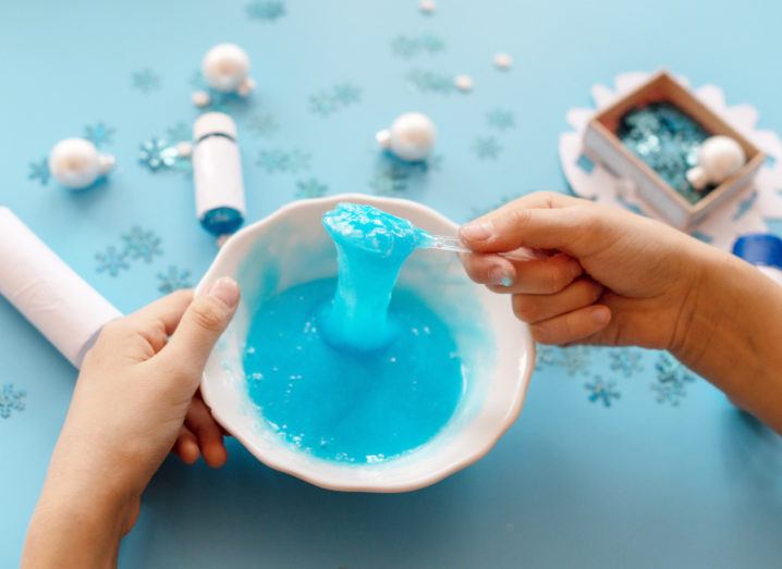 A child’s hands holding a bowl and spoon with blue slime in it, on a light blue table with snowflake confetti and glitter scattered around.