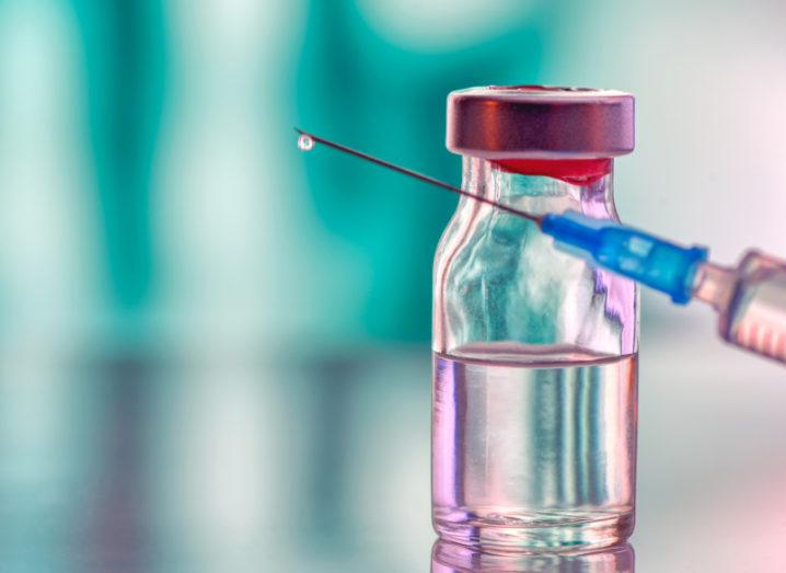 A close up of a medicine vial with a vaccine sitting on a table, while a syringe is held at an angle in front of it.