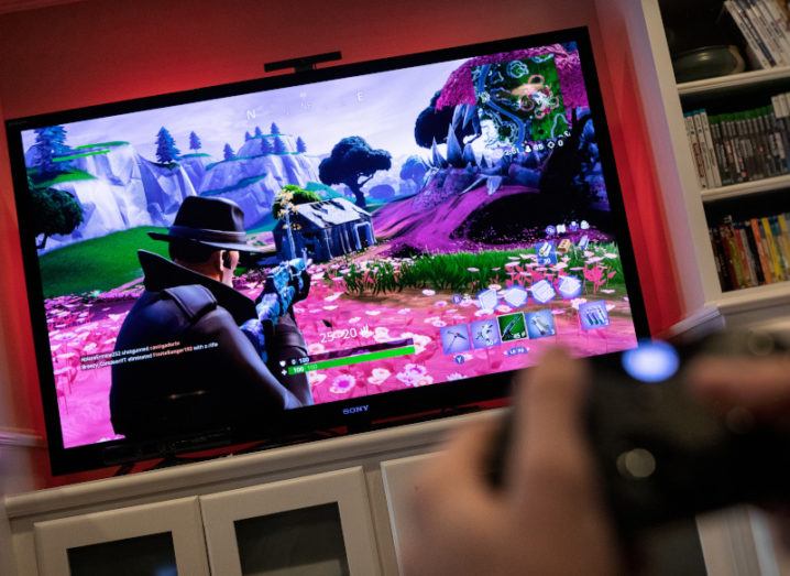 View from a game controller at a large TV screen in a living room showing a video game in play, with a character pointing a gun at a shack in a colourful mountain landscape.