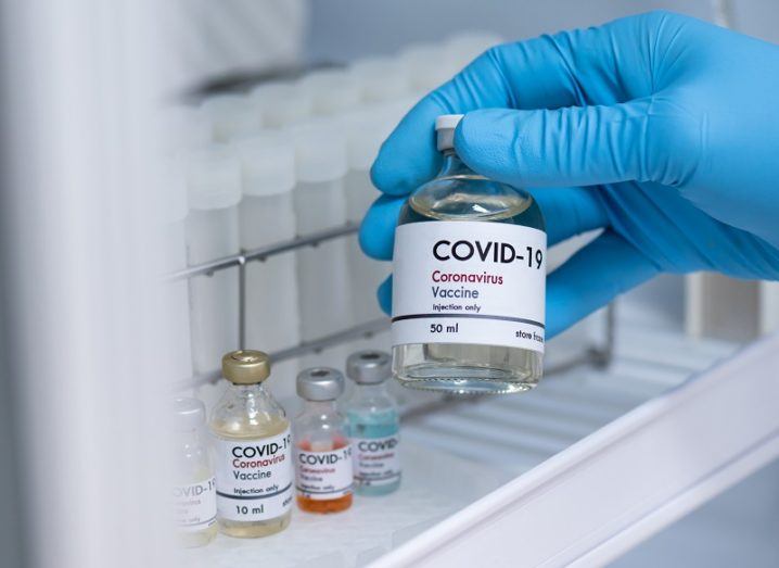 A gloved hand holding a vial of a Covid-19 vaccine in a refrigerator.