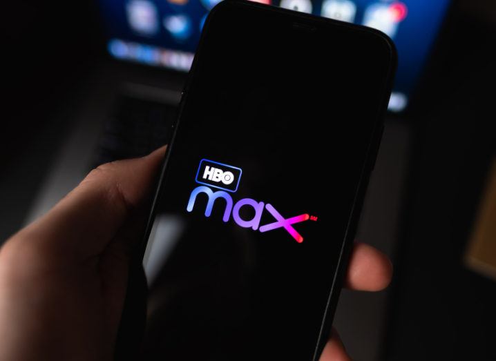 A hand holding a smartphone displaying the HBO Max logo. The person is a darkened living room, preparing to stream a film.