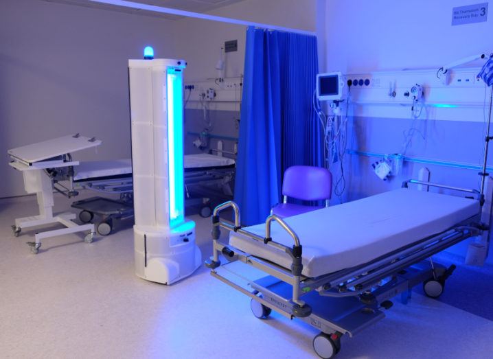 A tall robot beams an ultraviolet light into a hospital ward containing beds and other medical equipment.