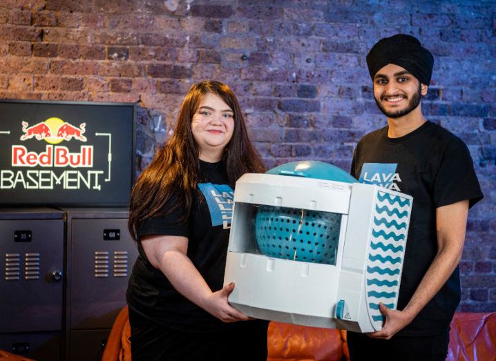 A young woman and young man stand beside a sign that says Red Bull Basement while holding a large white box, which is a portable washing machine.