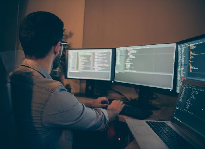 The back of a man sitting at a desk with three large monitors and a laptop in front of him, all displaying lines of code.