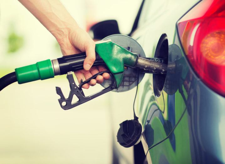 A hand clasped around a green fuel pump, which is inserted into the tank of a blue car.