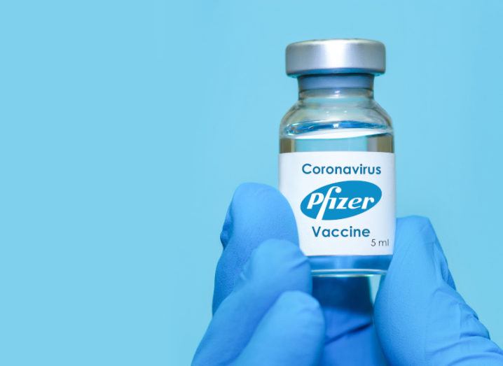 A hand wearing blue surgical gloves holds a small vial with Pfizer Covid-19 vaccine in it and labelled as such against a bright blue background.