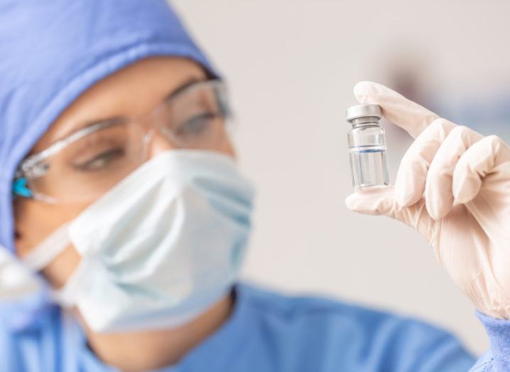 A healthcare worker in protective equipment is holding up a vial of a vaccine.