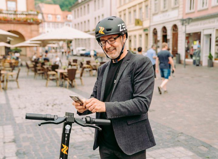 Damian Young, CEO of Zeus Scooters, is standing on a street with a scooter and an app.
