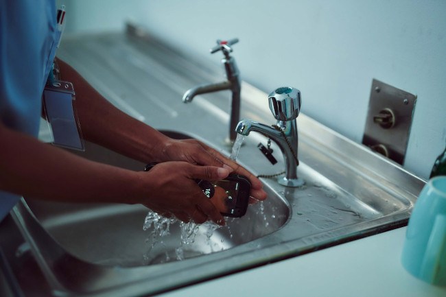 A woman’s hands washing a Cat smartphone in a sink under a running tap. 