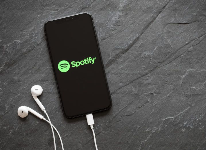 A mobile phone with the Spotify logo on its screen is lying on a grey surface with white earphones.