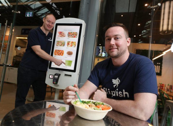 Flipdish founders Conor and James McCarthy. Conor stands at digital food-ordering kiosk while James sits at a table with a plate of food.
