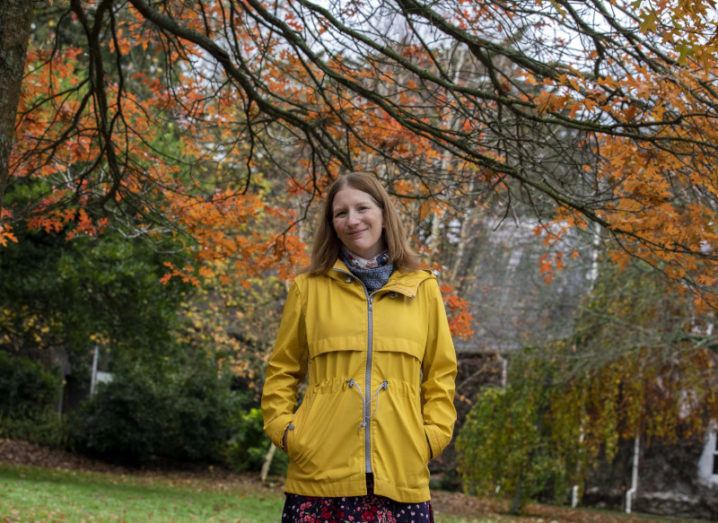 A woman in a floral dress and yellow rain jacket stands with her hands in her pockets in a park where nearby trees’ leaves have turned burnt orange.