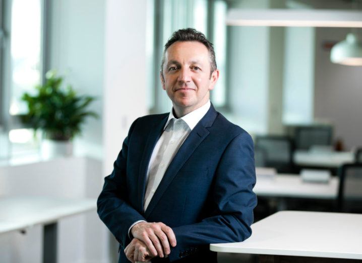 Steve Coakley of BT Ireland leans against a white surface while smiling at the camera. He is dressed in a business suit.