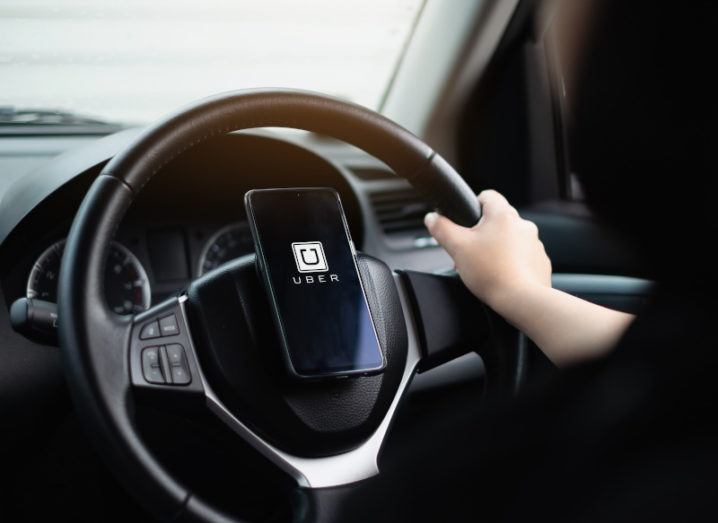 The inside of a car, looking at the steering wheel from behind. There is one hand on the wheel and a phone attached to the centre with an Uber logo on the screen.