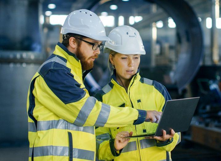 Two engineers in protective clothing stand on-site looking at a laptop.