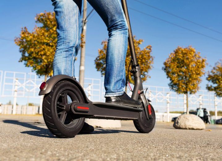 The legs of a man standing on an e-scooter parked on a road against a bright blue sky.