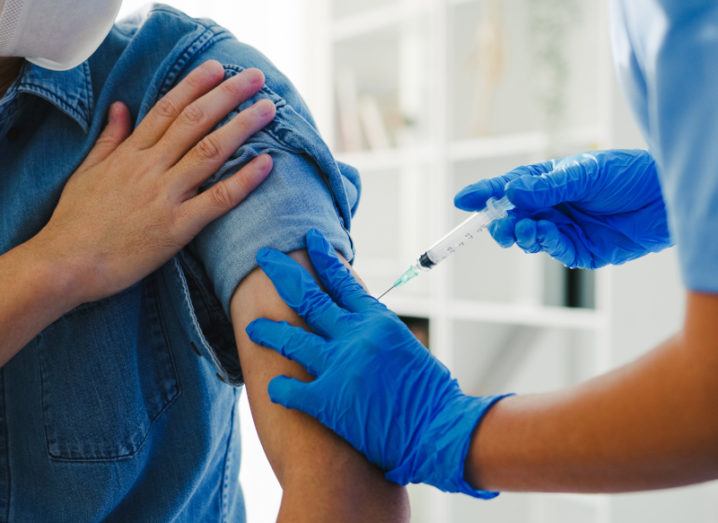 A nurse in protective clothing is giving a vaccine shot to a young man in a clinical setting.