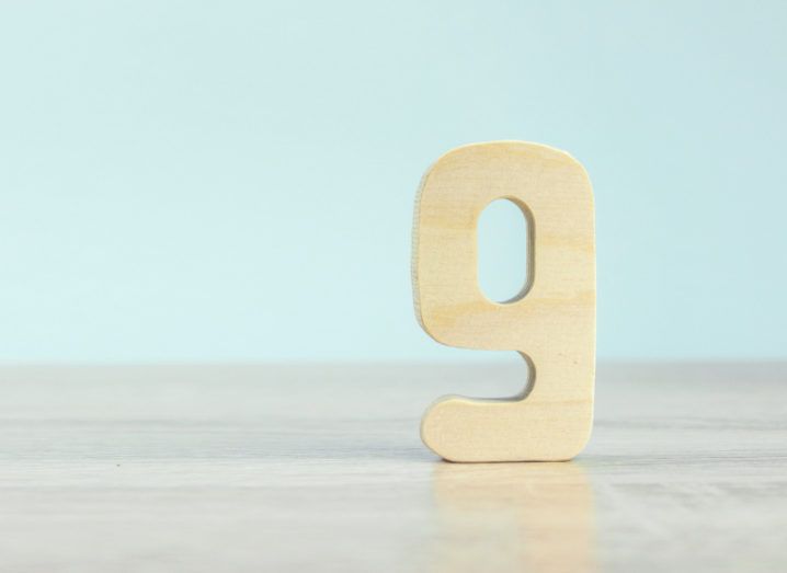 A wooden number 9 standing upright on a wooden table against a baby blue wall.
