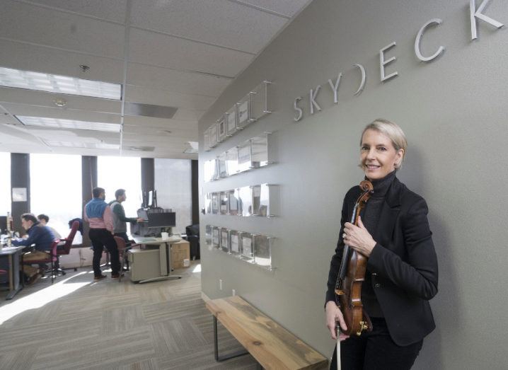 A woman holding a violin leans against an office wall that says 'SkyDeck'.